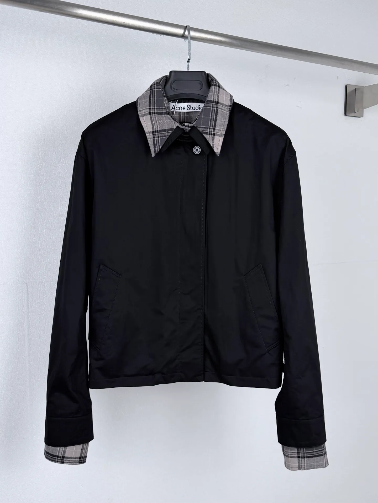 ACNE Studios black plaid collar fake two-piece short lapel jacket relies on double-layer cutting to create the visual effect of layering shirts. The difference in fabric properties and the superposition of multiple splicing points make the entire production process have a high process threshold and a very low production error tolerance rate. The main body of the garment is made of washed twill woven fabric, and the splicing parts are matched with Retro plaid fabric. The two types of fabrics have different weights and shrinkage rates, and pre-shrinking and shaping processes must be performed separately before production. If the fabric pre-treatment is not in place, the shrinkage of the two fabrics will be inconsistent after the garment is completed, and the spliced ​​seams will easily appear to be pulled, wrinkled, and twisted. The lapel is composed of a solid color base fabric and a plaid pattern. During the sewing process, it is necessary to continuously balance the tension of the two layers of fabrics and strictly control the collar angle arc to ensure that the left and right collar shapes are symmetrical and upright, and to prevent blistering and folding and misalignment of the collar surface. The checkered cuff cuffs are made of independent split pieces. The direction of the checkered pattern is calibrated during the cutting stage. The textures on the left and right cuffs are in the same position, and the splicing horizontal lines are consistent to maintain a coordinated appearance. The front panel adopts a hidden placket structure, with multiple layers of overlapping stitching and obvious thickness differences. The sewing pressure and stitch length are dynamically adjusted during sewing to avoid problems such as skipped stitches and wavy seams. Adjustment tab components are configured on both sides of the hem, and the positioning accuracy of the small auxiliary pieces is stringent. The diagonal pocket adopts embedded technology, and the bag fabric is seamlessly connected with the body without destroying the simple lines of the body. The vertical dividing line of the garment body is precisely aligned up and down to ensure the square shape of the short box-shaped silhouette and prevent the left and right asymmetry of the garment and the height deviation of the hem. Difficulties such as splicing different materials, multi-layer composite sewing, and alignment of plaid parts are superimposed on each other. Improper tension control in any process will weaken the natural texture of two fake pieces. From fabric pre-treatment, precise cutting, layered precision sewing, to low-temperature flexible ironing and shaping, the process chain is complicated, testing the lathe craftsmanship and the factory's refined quality control capabilities, and clearly reflecting the manufacturing process barriers of light luxury garments. Size：36 38 40 42
